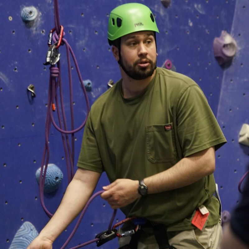 The image shows a man wearing a green helmet and shirt, standing in front of a climbing wall. He appears to be holding a rope, suggesting he is either preparing to climb or assisting someone else. The background includes climbing holds and ropes, indicating an indoor climbing gym.